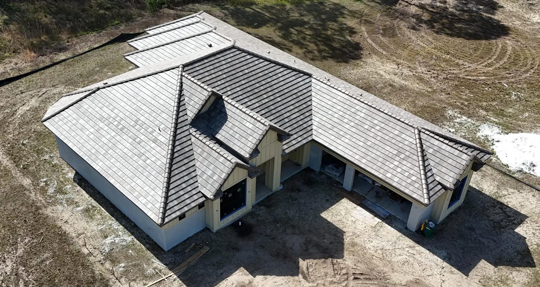 Aerial view of a new home under construction with a gray tile roof, surrounded by bare dirt lot and wooded area